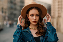 Outdoor Close Up Fashion Portrait Of Young Elegant Lady Wearing Beige Fedora Hat, Trendy Chain Necklace, Blue Denim Shirt, Posing In Street Of European City. Copy, Empty Space For Text
