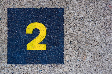 Yellow number 2, two, in dark blue square, offset on flat concrete surface.