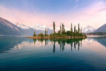 Battleship Island with Pine Trees reflected in calm water of Garibaldi Lake Landscape and Distant Snowcapped Peaks obscured by smoke and ash of summer Wilfdfires in Coast Mountains of British Columbia