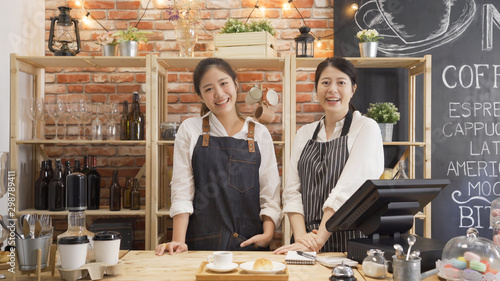 Portrait of cheerful asian female barista laughing during working break together with positive smile. Partnership of youg women in common business cafeteria. two girl staff face camera in coffee shop