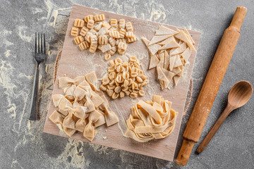 Various of freshly made homemade pasta on a cutting board, fork, rolling pin and wooden spoon on the kitchen table. Top view .