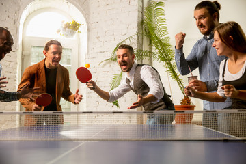 Wall Mural - Young people playing table tennis in workplace, having fun. Friends in casual clothes play ping pong together at sunny day. Concept of leisure activity, sport, friendship, teambuilding, teamwork.