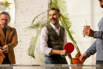 Wall Mural - Young people playing table tennis in workplace, having fun. Friends in casual clothes play ping pong together at sunny day. Concept of leisure activity, sport, friendship, teambuilding, teamwork.