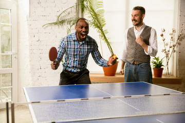 Wall Mural - Young men playing table tennis in workplace, having fun. Friends in casual clothes play ping pong together at sunny day. Concept of leisure activity, sport, friendship, teambuilding, teamwork.