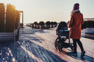 Wall Mural - Young woman walking through city park with baby stroller. Warm autumn or spring weather for outdoor activity. Pretty urban architecture and landscape design. Sunset, clouds in sky, sunny day for walk