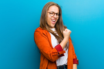 Wall Mural - young woman feeling happy, satisfied and powerful, flexing fit and muscular biceps, looking strong after the gym against blue background