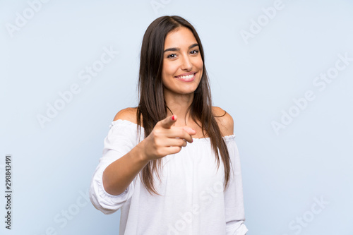 Fotomural Young woman over isolated blue background points finger at you