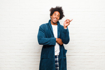 Wall Mural - young black man wearing pajamas with gown smiling happily and looking sideways, wondering, thinking or having an idea against brick wall