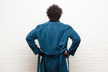 Wall Mural - young black man wearing pajamas with gown feeling confused or full or doubts and questions, wondering, with hands on hips, rear view against brick wall