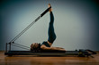 © Georgii - Young girl doing pilates exercises with a reformer bed. Beautiful slim fitness trainer on a reformer gray background, low key, art light, copy space advertising banner