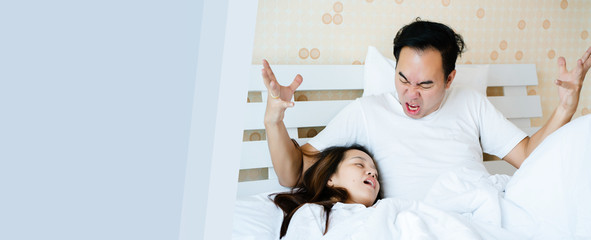 Asian women are snoring with loud noises, creating angry and annoyance for men. Couple lying on the white bed. Snoring may lead to apnea and death. Health care concept. Banner for text copy space