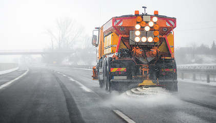 Snow plow salting street in winter time. Orange truck deicing. Maintenance winter vehicle back side.