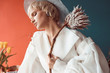 © monchak - portrait of beautiful girl with flower in hands in front of turquoise and orange wall, she looks down, she is dressed in white dress and hat on the head and she has professional nude makeup