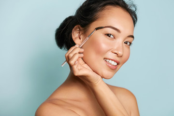 Wall Mural - beauty. smiling woman brushing eyebrows with makeup brush closeup. portrait of beautiful happy asian