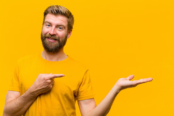 young blonde man smiling, feeling happy, carefree and satisfied, pointing to concept or idea on copy space on the side against orange wall