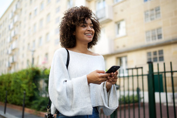 Wall Mural - smiling young african american woman walking in city with mobile phone in hand
