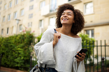 Wall Mural - portrait of smiling young african american woman walking in city with cellphone in hand
