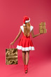 back view of young african american girl in santa hat and christmas dress holding gift boxes and shopping bag on red background