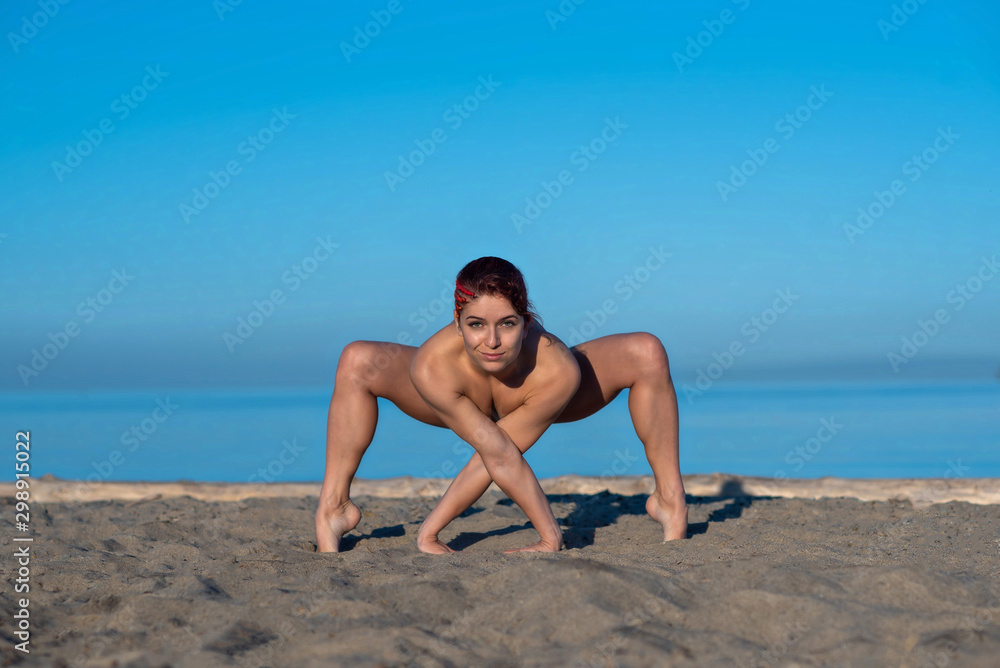 A beautiful nude woman practices yoga alone on the seashore. Red-haired girl  is engaged in Pilates without clothes by the sea. Clear sky. pacification  and zen. Very flexible athlete. Stock Photo |