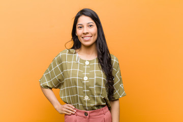 young pretty hispanic woman smiling happily with a hand on hip and confident, positive, proud and friendly attitude against brown wall