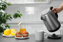 Woman Pouring Water From Modern Electric Kettle Into Cup At Grey Table In Kitchen, Closeup