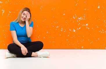 Wall Mural - young blonde woman laughing and slapping forehead like saying d‚Äôoh! I forgot or that was a stupid mistake sitting on the floor