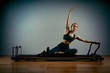 © Georgii - Young girl doing pilates exercises with a reformer bed. Beautiful slim fitness trainer on a reformer gray background, low key, art light, copy space advertising banner