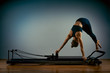 © Georgii - Young girl doing pilates exercises with a reformer bed. Beautiful slim fitness trainer on a reformer gray background, low key, art light, copy space advertising banner