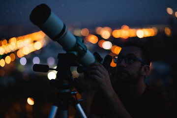 Wall Mural - astronomer with a telescope watching at the stars and moon with blurred city lights in the backgroun