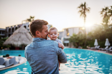 Handsome Caucasian dad hugging son and looking at him while standing next to swimming pool. Baby looking over father's shoulder.