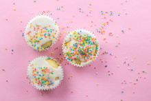 Three Little White Frosted White Cupcakes In Centre Of Pink Background Sprinkled With Multi - Coloured Sprinkles On Pink With Copy Space - Top View