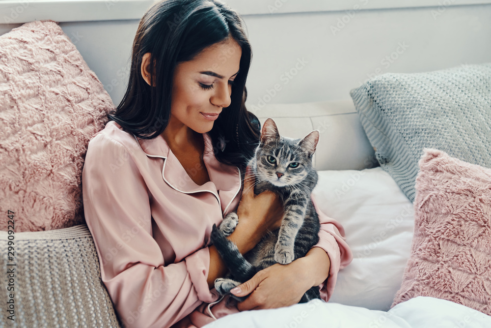 Beautiful young woman in pajamas smiling and embracing domestic cat while resting in bed at home - obrazy, fototapety, plakaty 