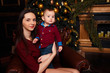 © Skripnik Olga - Mom sits on a chair with a baby in her arms, hugs and kisses. Christmas gift, family and happiness, Christmas tree and holiday