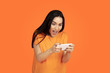 © master1305 - Caucasian young woman's portrait on orange studio background. Beautiful female brunette model in shirt. Concept of human emotions, facial expression, sales, ad. Copyspace. Plays smartphone, win in bet