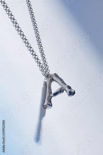 Object photo of a silver letter A pendant on on a chain, lying on a blue background. The pendant is casting a shadow.