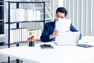 A young businessman sitting in a modern office. He has a feel stressed about the result of business profits not positive. On his table have a computer laptop tablet pen paper graph.