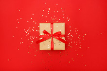 Christmas Composition Greeting Card. Gift From Craft Paper On A Red Background With A Gold Star Confetti. Top View, Flat Lay.