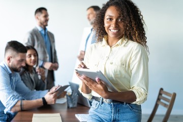 Sticker - Beautiful young professional african woman standing in office while workers hold a meeting in background.