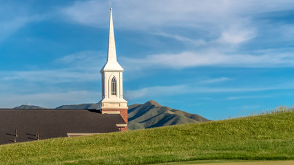 Wall Mural - Panorama Roof and steeple of a church viewed from a golf course against cloudy blue sky