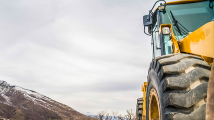 Wall Mural - Panorama Close up of the tires of a bulldozer against snow peaked mountain and cloudy sky