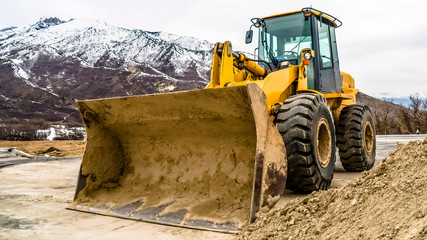 Wall Mural - Panorama Front view of a yellow bulldozer against snow topped mountain and cloudy sky