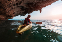 Woman Paddles Kayak In The Tropical Sea At Sunset And Passes The Limestone Mountains With Stalactites. Model Looks At The Camera And Smiles