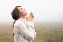 Girl Closed Her Eyes, Praying In A Field During Beautiful Fog. Hands Folded In Prayer Concept For Faith