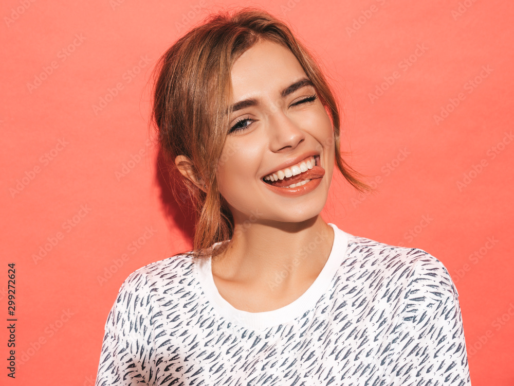 Young beautiful woman looking at camera. Trendy girl in casual summer shirt clothes with natural makeup. Positive female smiling. Funny model posing near pink wall in studio.Shows tongue and winks - obrazy, fototapety, plakaty 