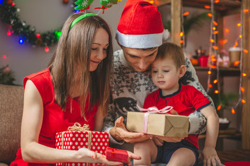 Wall Mural - Mom and dad give their son a gift in a red box. A young family with a child celebrates Christmas and New year at home