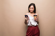 © dianagrytsku - Full length portrait of a smiling asian woman using mobile phone while holding cup of coffee to go isolated over beige background