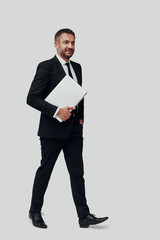 Full length of charming young man in full suit carrying laptop and smiling while walking against grey background