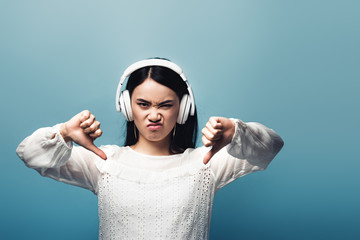 Wall Mural - angry asian woman with headphones showing thumbs down on blue background