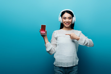 Wall Mural - smiling asian woman with headphones pointing with finger at smartphone with trading courses app on blue background