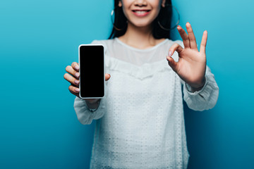 Wall Mural - cropped view of smiling asian woman in white blouse showing ok sign and smartphone with blank screen on blue background